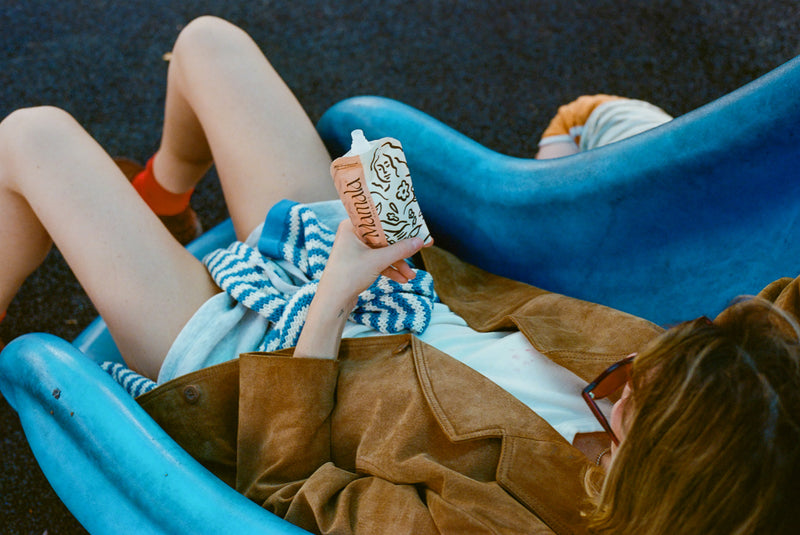 Person reclining on a blue lounge chair, holding a decorated drink carton and wearing sunglasses and a brown jacket.