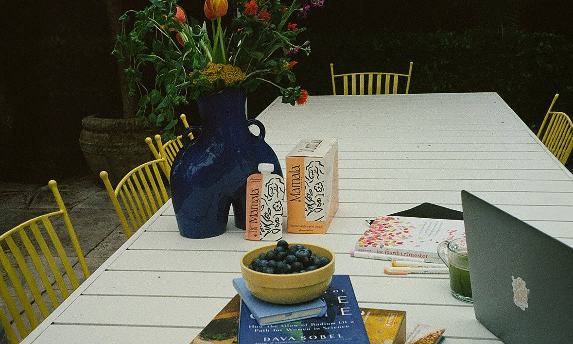 Mamala Organics pouch and packaging leaning against a flower vase on top of a white table, next to fresh fruit, books, and a laptop.