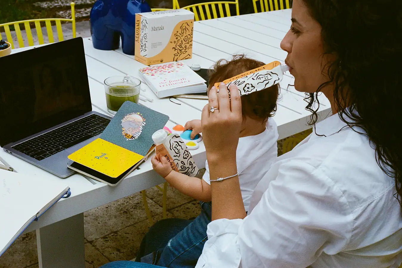 Woman in white shirt holding a baby on her lap, working on a laptop, and sipping from a Mamala Organics pouch.