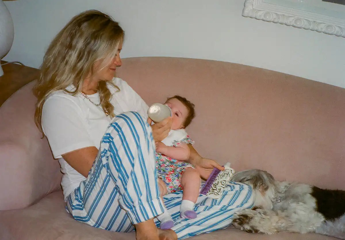 A woman feeding a baby from a bottle, sitting on a couch next to a dog, and holding a Mamala Organics pouch.