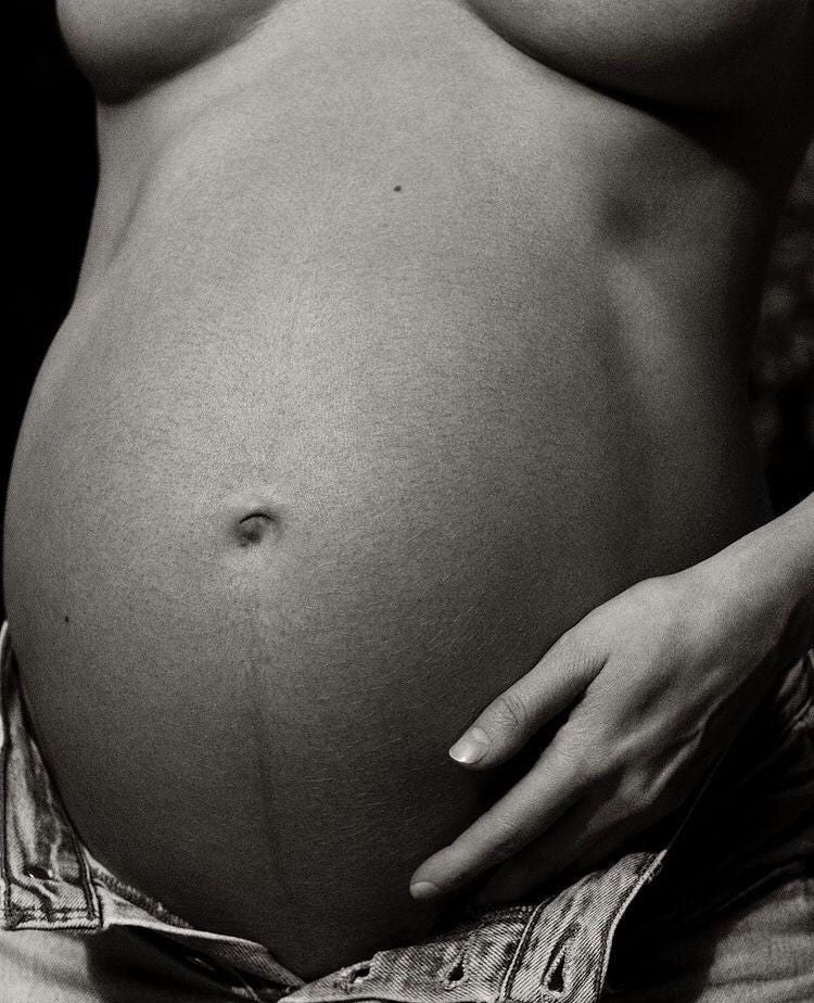 Pregnant woman gently holding her belly while preparing for a supported postpartum and fourth trimester.”