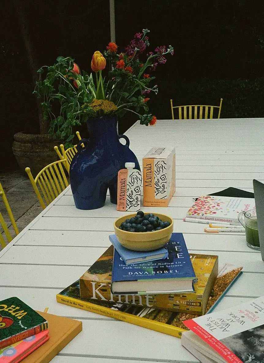 Table with books, a vase of flowers, a bowl of fruit, and Mamala Organics package and pouch on a white table outdoors.
