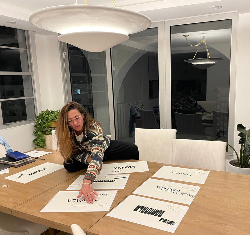 A woman with long light brown hair sits at a wooden dining table in a modern room, pointing at printed white cards with black text. Several cards are laid out on the table, including one that reads 'NADIRA' and another that reads 'MAMBA NADIRA'.