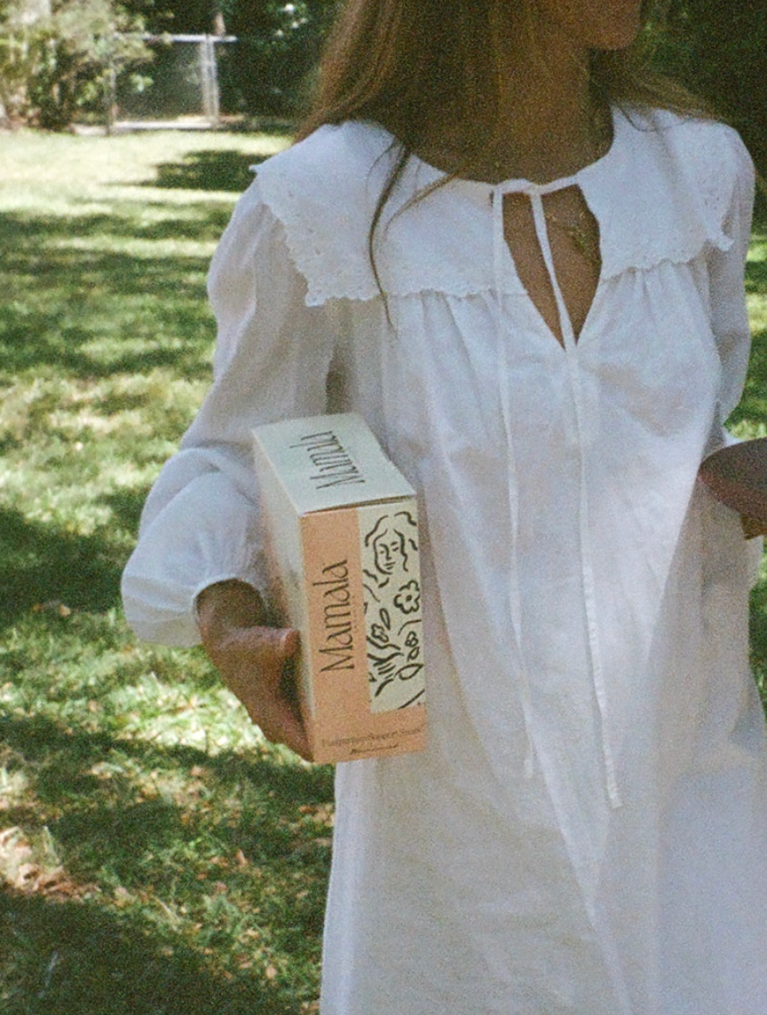A woman outdoors in a grassy area wearing a white long-sleeve nightgown or dress and holding a beige box labeled 'Mamala'.