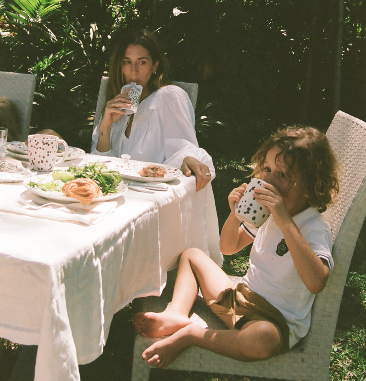A woman and a young child sitting in outdoor wicker chairs at a dining table, both holding speckled white mugs. The scene is set in a lush garden with a white tablecloth and breakfast dishes.