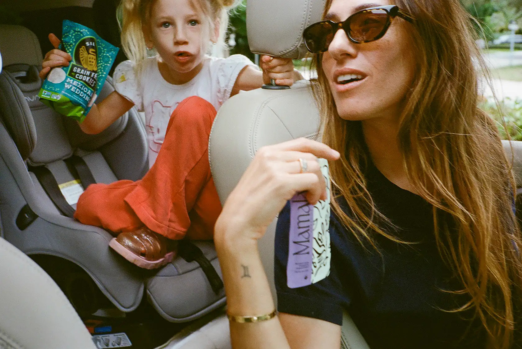 Woman driving a car, wearing sunglasses, and child in a car seat with snacks. Woman is holding Mamala Organics pouch.