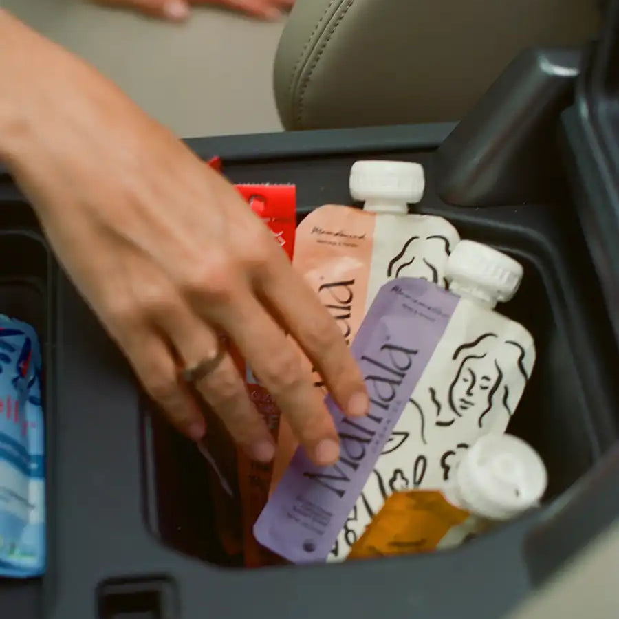 Hand reaching into a car console tray with various items including bottles and Mamala Organics pouches.