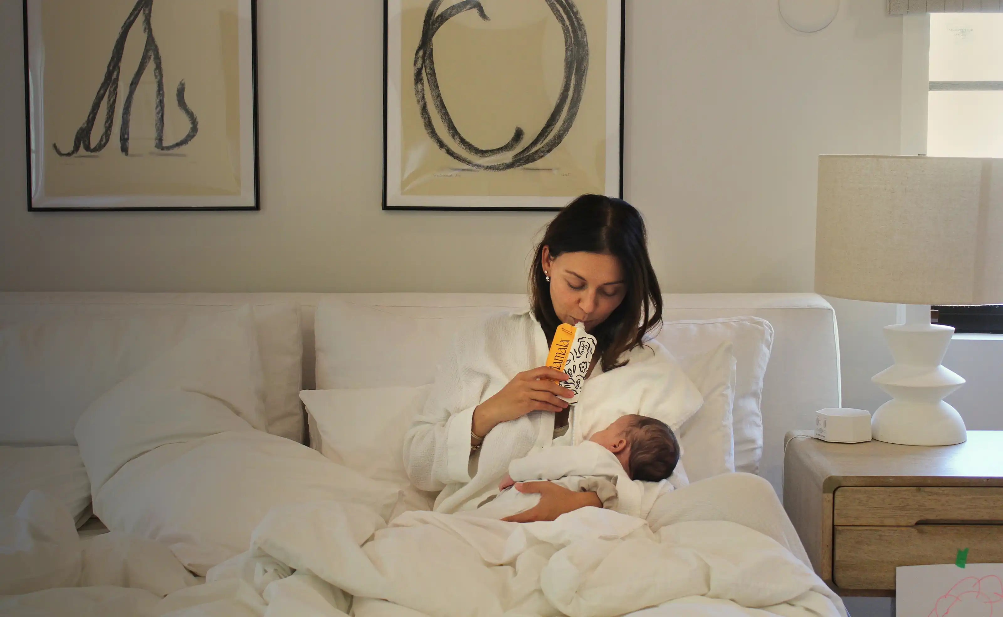 Woman in a white robe holding a baby in bed with a Mamala Organics pouch, in a bedroom setting.